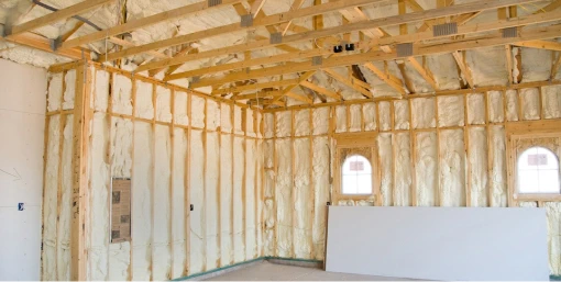 Bright interior of a building under construction, with exposed wooden framing and insulation. Walls and ceiling are covered with a layer of sprayed foam insulation, which fills the gaps between the wooden studs and rafters, creating a uniform insulated surface