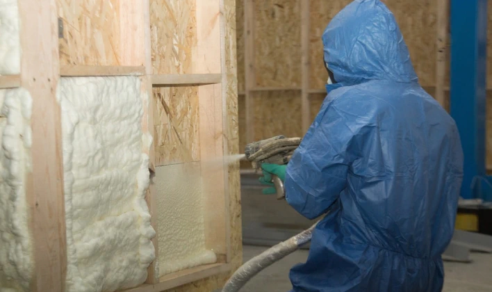 An experienced worker in a blue protective suit is spraying foam insulation into the wooden framework of a wall. The foam is expanding and filling the spaces between the wooden studs