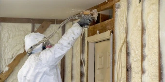 A worker in white protective gear sprays foam onto exposed wood studs and wall voids to fill them in providing effective thermal insulation